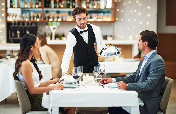 Professional waiter serving covered dish to couple dining at elegant white-tablecloth restaurant table