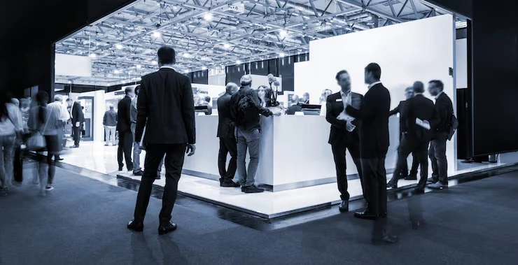 Business professionals networking around modern white exhibition booth in large convention hall with geometric ceiling