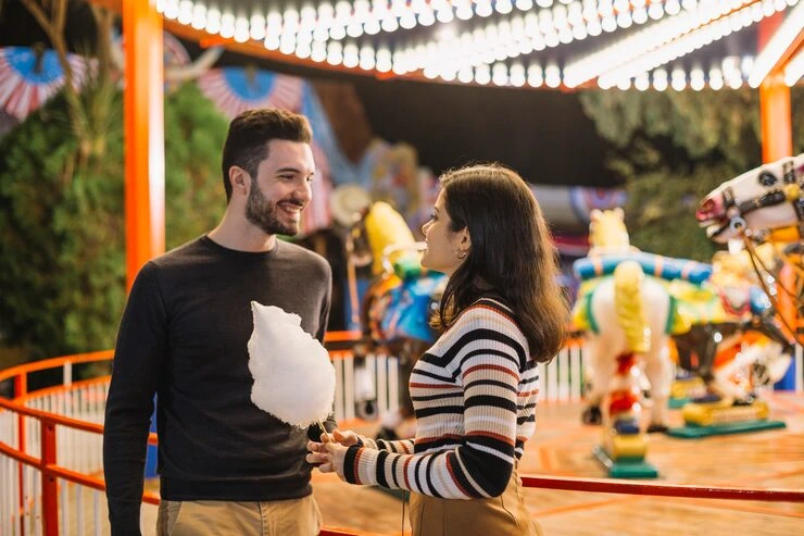 Couple enjoying carnival atmosphere with cotton candy under colorful string lights and amusement rides