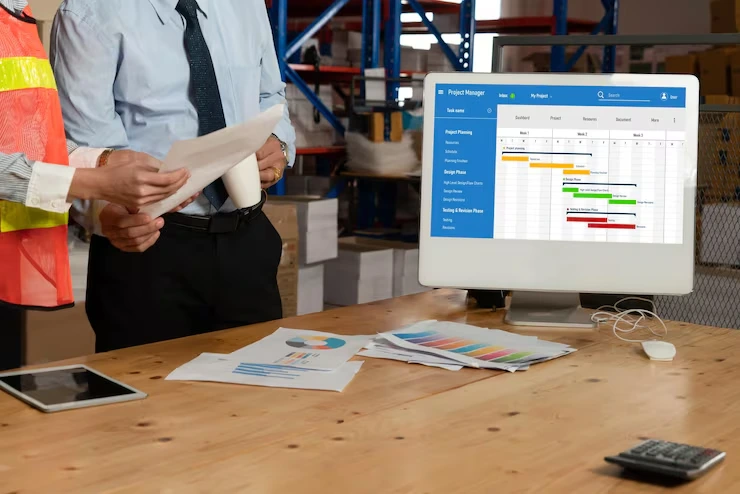 Manager reviewing project timeline charts on computer monitor while warehouse supervisor holds documents in logistics facility