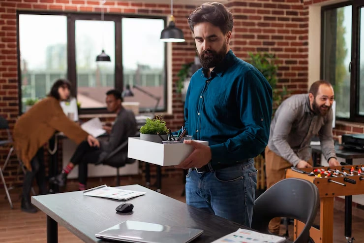 Professional carrying box with plant and supplies while colleagues work in modern brick office with large windows
