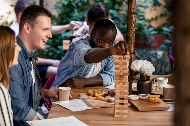 Colleagues playing wooden block tower game while enjoying coffee and snacks at outdoor cafe table