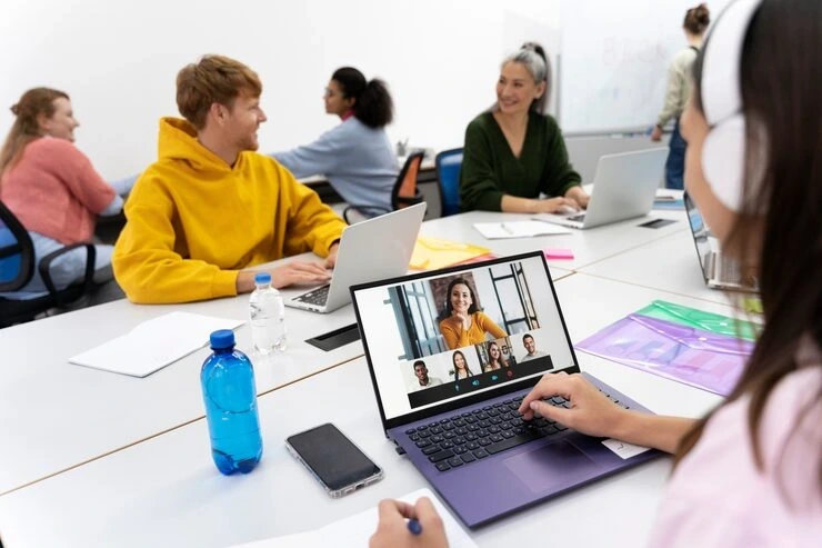 Team members working on laptops with video conference call displayed while colleagues collaborate in background